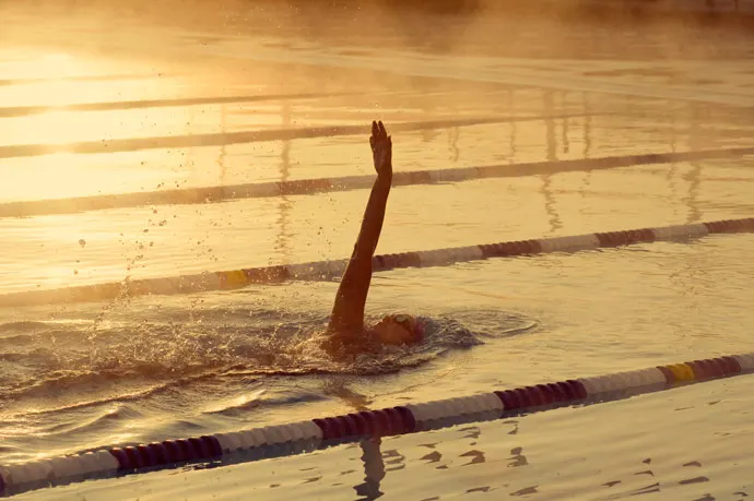 On a sunny morning, a young man swims backstroke in an outdoor pool while steam rises from the water.
