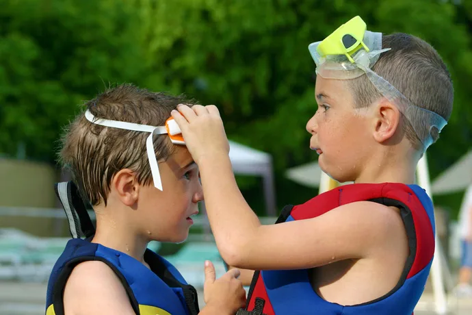 A young boy helps another young boy to put on swimming goggles.